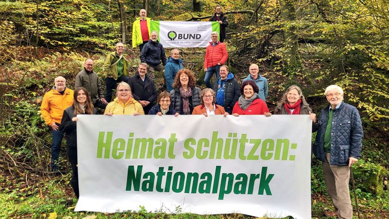 Viele Menschen halten ein Banner mit der Aufschrift „Heimat schützen: Nationalpark“. Im Hintergrund ist ein BUND-Plakat zu sehen.