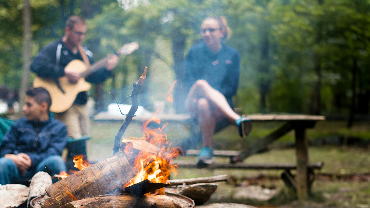 Jugendliche sitzen mit Gruppenleiter, der Gitarre spielt, um ein Lagerfeuer.