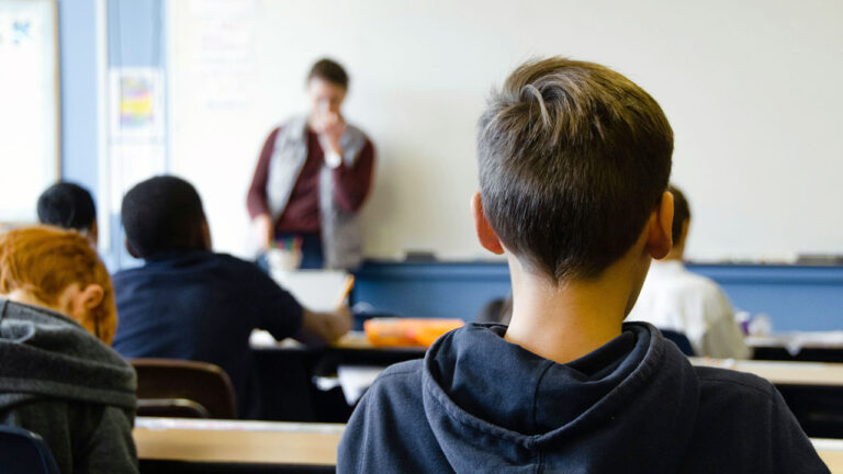 Von hinten aufgenommene Kinder sitzen in einer Schulklasse vor einem Erwachsenen.