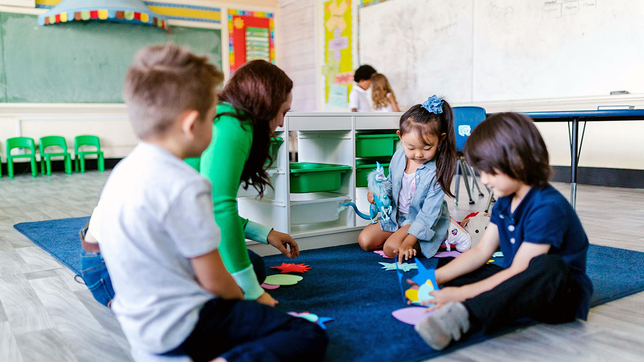 Mehrere Kinder sitzen in einem Klassenraum gemeinsam mit einer Erwachsenen auf einem blauen Teppich und spielen mit geometrischen Formen.