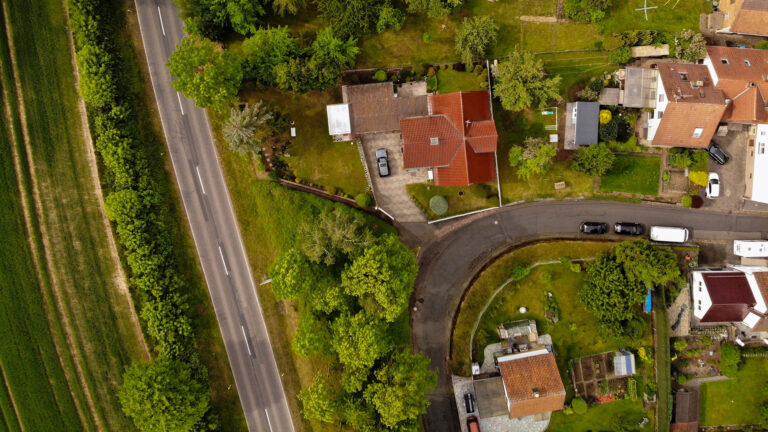 Drohnenbild einer Ansammlung von mehreren Häusern, Straßen, Autos und Wiesen in der Aufsicht.