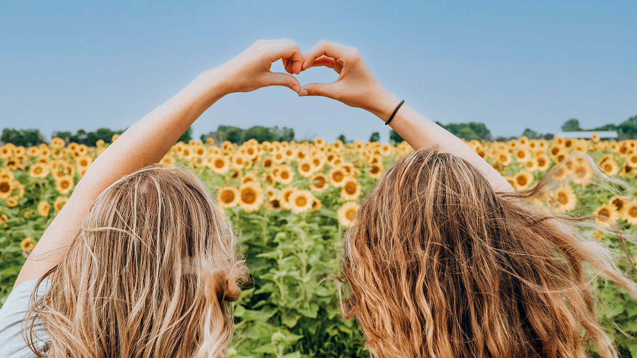 Zwei von hinten aufgenommene Frauen vor einem Sonnenblumenfeld, die mit ihren Händen ein Herzsymbol formen.